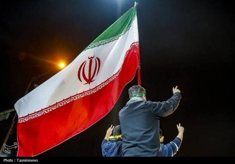 At night, a man sits on another's shoulders waving a large Iranian flag toward a stadium crowd.