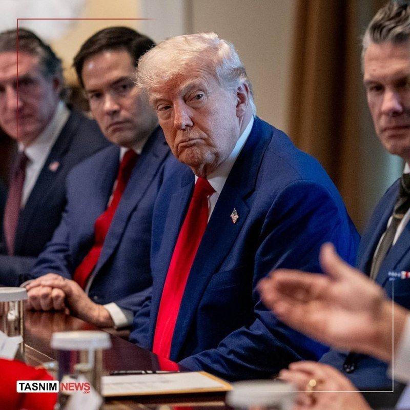 I can't identify the person based on their appearance. I can describe what I see: a blonde man in a dark blue suit and red tie gestures with both hands raised while speaking, with people and flags visible behind him.