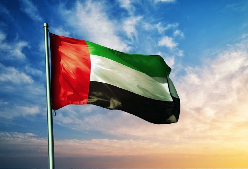 The Iranian flag and Russian flag wave side by side on a flagpole against a blue, partly cloudy sky.