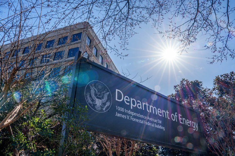 A sign reading "Department of Energy, 1000 Independence Avenue SW, James V. Forrestal Federal Building" stands before a modern office building under a bright sun.
