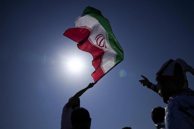 A bespectacled man in a dark suit stands at an ornate wooden podium bearing Persian script and the Iranian emblem, flanked by two Iranian flags against a backdrop displaying a world map and "Ministry of Foreign Affairs."