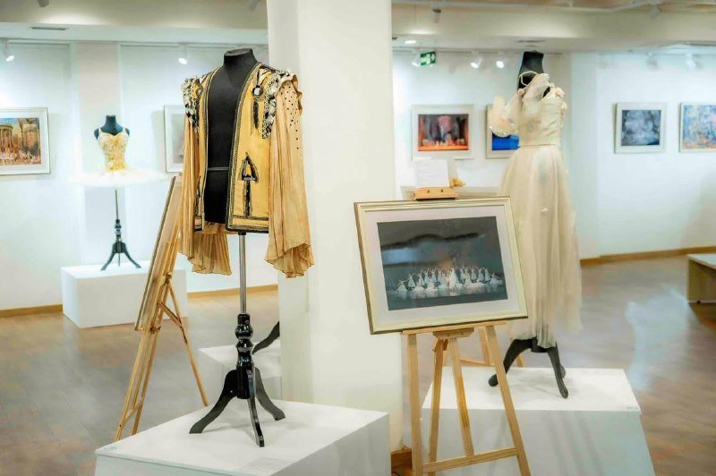 A gallery displays embroidered garments on mannequins alongside framed paintings and a ballet photograph mounted on easels.