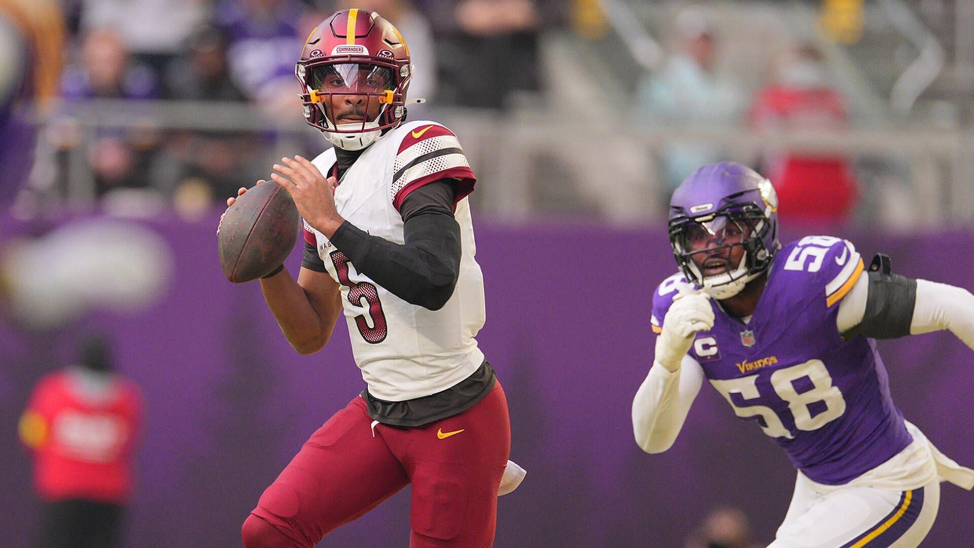 A football player in a white and burgundy jersey scrambles with the ball while a defender in a purple Vikings jersey number 58 pursues during a game.