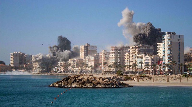 Thick plumes of dark gray smoke rise from explosions among beachfront high-rise buildings along a coastal city, with birds flying nearby and calm blue water in the foreground.