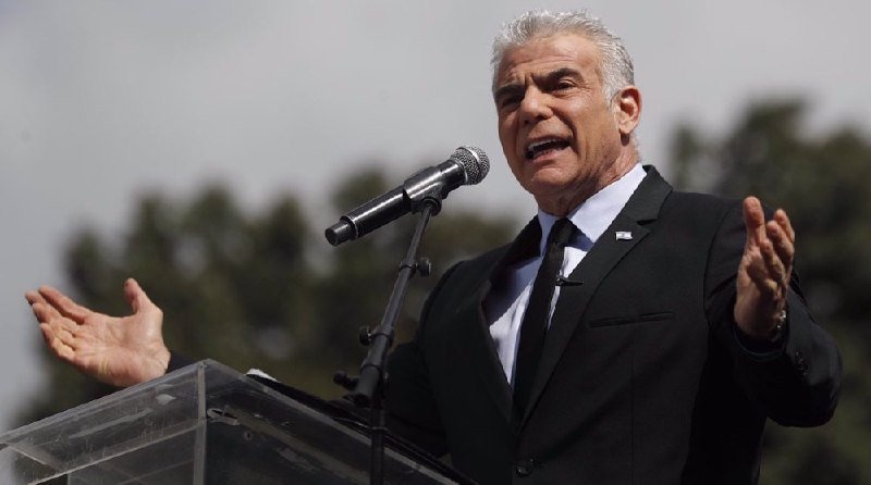 A gray-haired man in a dark suit and tie speaks into a microphone at a clear podium, gesturing with both hands raised against a blurred outdoor background of trees.