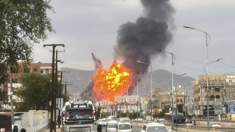 A large orange fireball with a thick black smoke plume erupts in an urban street, with vehicles stopped nearby.