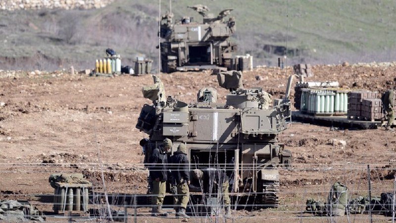Two armored military vehicles and soldiers in green uniforms are positioned in a rocky, barren field.