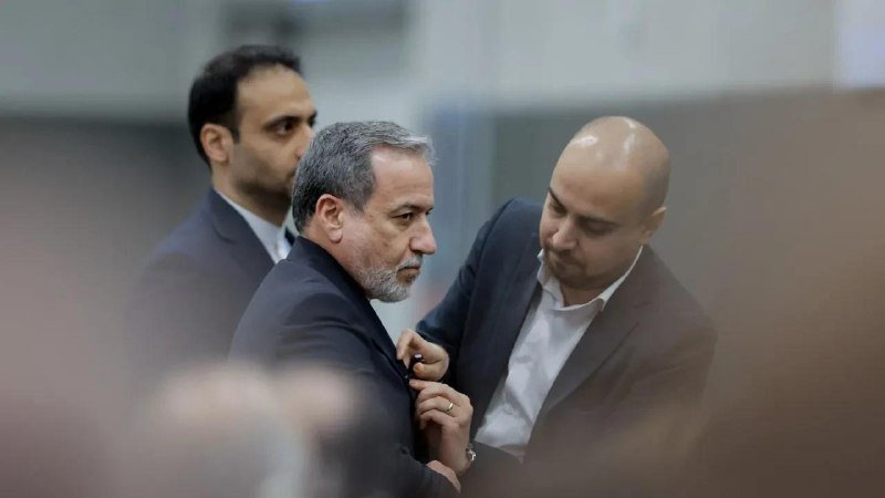 A gray-bearded man in a dark suit speaks with a bald man in a black suit and white shirt in a plain indoor setting.
