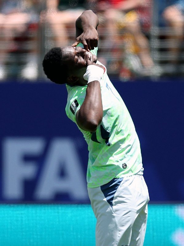 A soccer player wearing a red jersey with the number 7 and purple shorts celebrates with a raised fist on a stadium field.