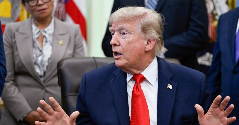 A blond man in a navy suit and blue tie speaks while seated, with an American flag and other people visible in the blurred background.