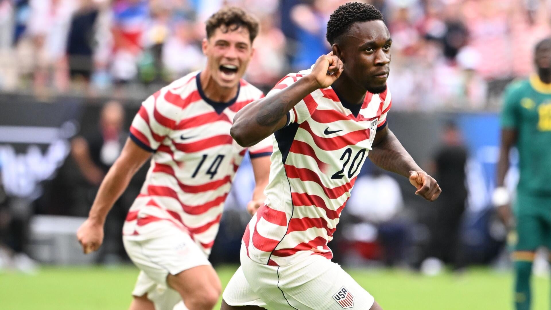 Two soccer players compete for the ball during a match, with the USA player in a dark jersey shielding it from an opponent in white.