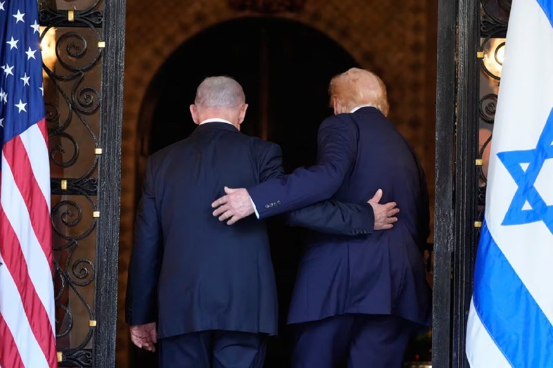 Two men in dark suits walk through an ornate doorway, arms around each other, flanked by an American flag and an Israeli flag.