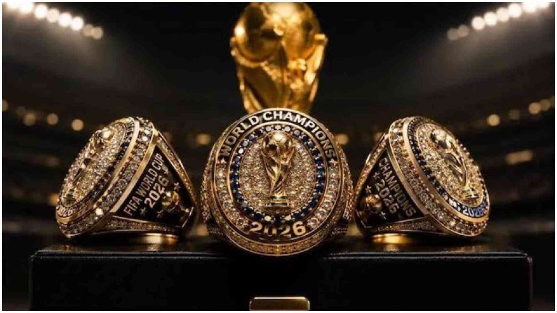 Three gold "FIFA World Cup Champions 2026" championship rings rest in front of a golden World Cup trophy against a blurred stadium backdrop.