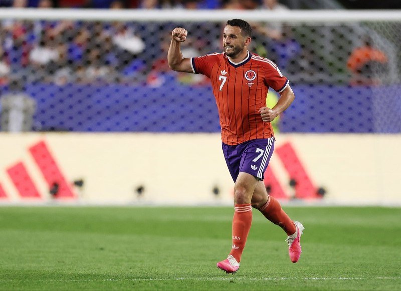 A soccer player in a red and purple uniform with the number 7 raises his fist in celebration while running on a green field, with a blurred crowd in the background.