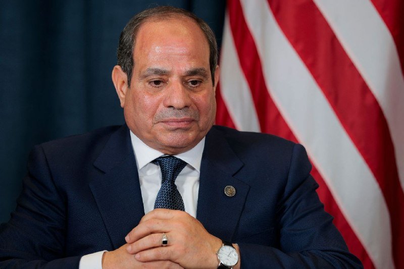 A man in a dark suit, white shirt, and patterned blue tie sits with hands clasped, an American flag visible behind him.