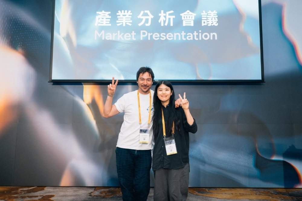 Two people wearing lanyards pose with peace signs in front of a screen displaying "Market Presentation" in English and Chinese.