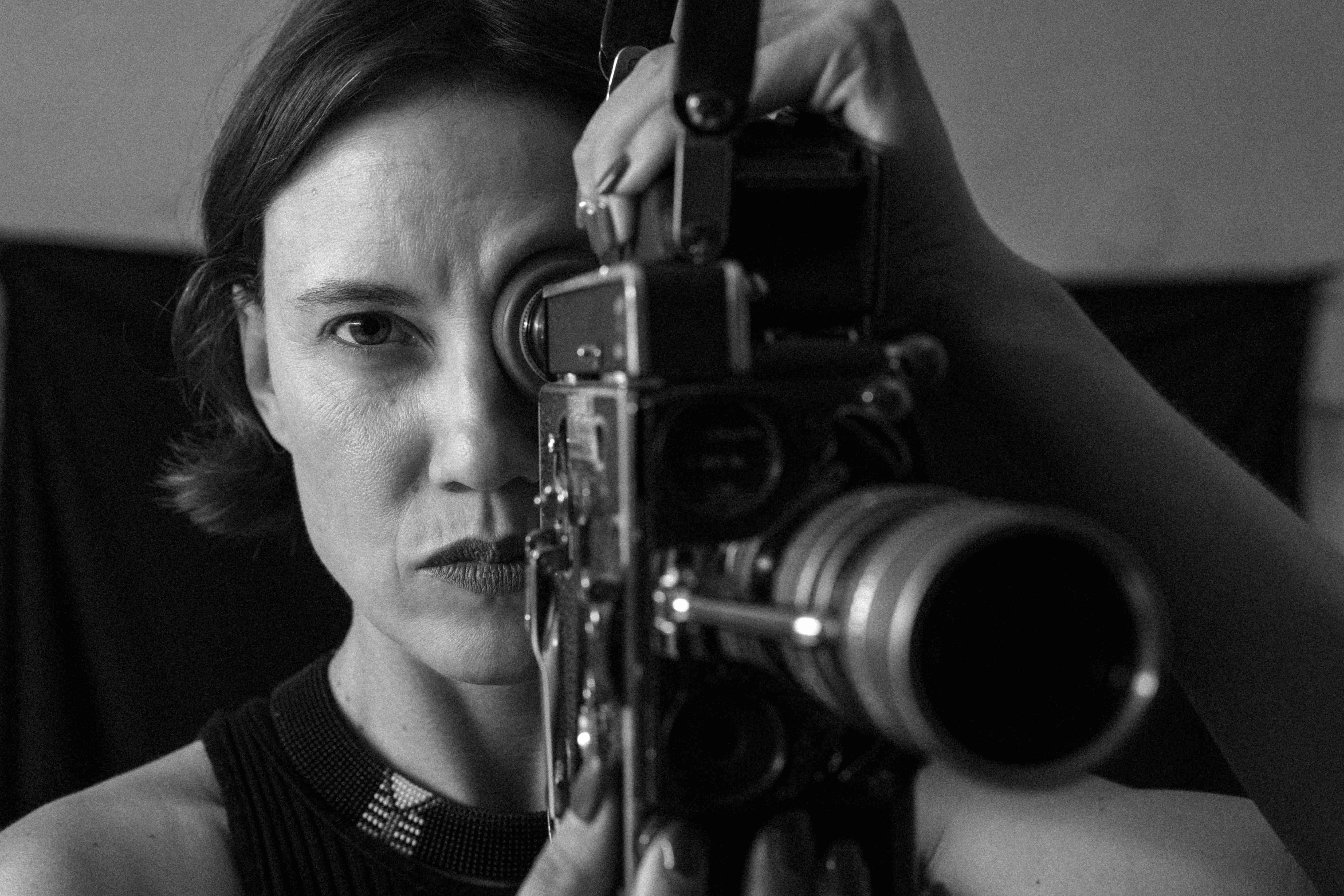 Black-and-white close-up of a woman looking through the viewfinder of a vintage movie camera, her hand adjusting its top mechanism.