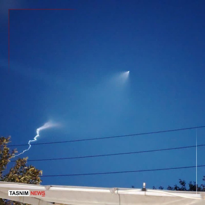 A bright object with a glowing trail streaks across a clear blue sky, leaving a white smoke plume behind power lines and tree tops.