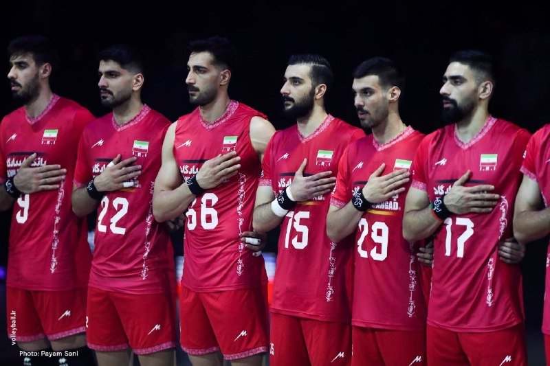 Seven men in red sleeveless jerseys with Iranian flag patches stand in a line with their hands over their hearts during what appears to be a national anthem ceremony.
