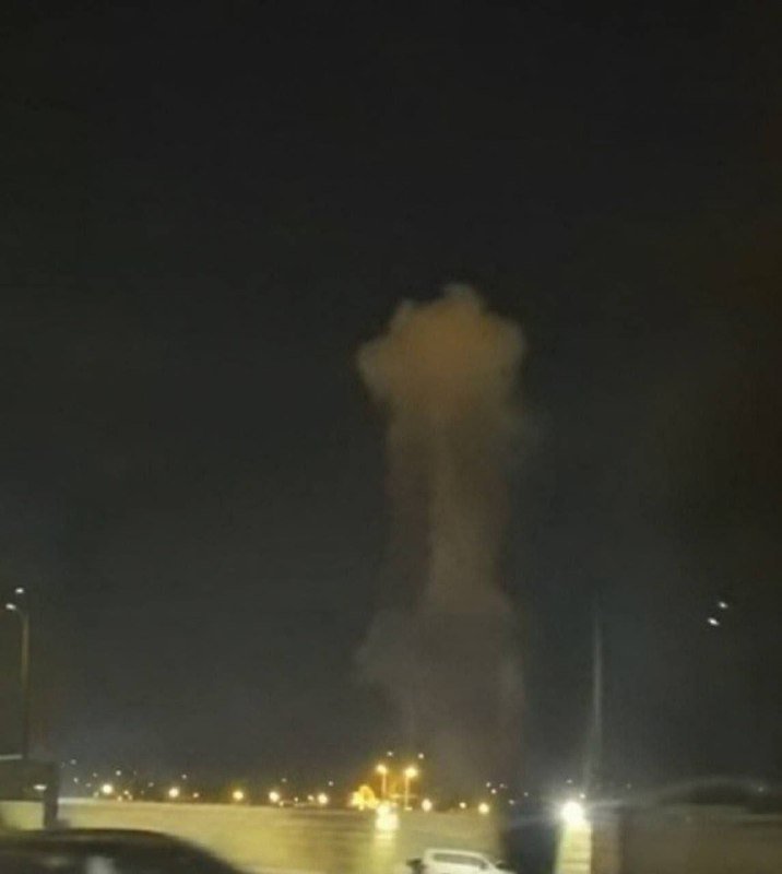 A blurry nighttime photograph shows a large plume of smoke or fire rising into a dark sky above a lit road with vehicles.