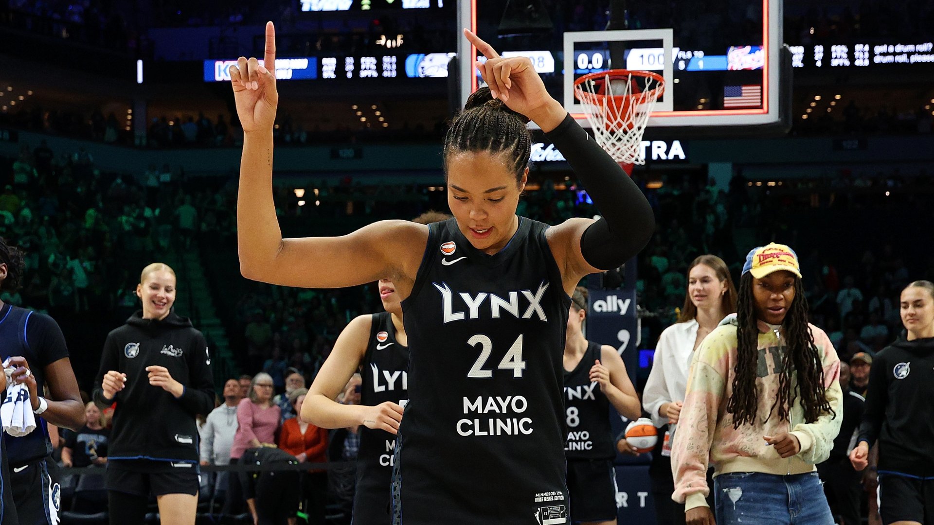 A Minnesota Lynx player wearing jersey number 24 raises both index fingers skyward on a basketball court, surrounded by teammates in black uniforms and a crowd in the background.
