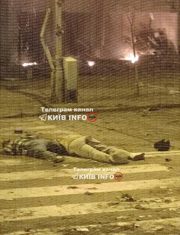 A person lies motionless on a debris-strewn street near a crosswalk at night, with smoke and damaged surroundings visible in the background.