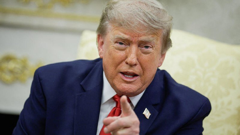 A man with blonde hair wearing a navy suit and red tie speaks while pointing a finger, seated in an ornate room with gold-trimmed decor.