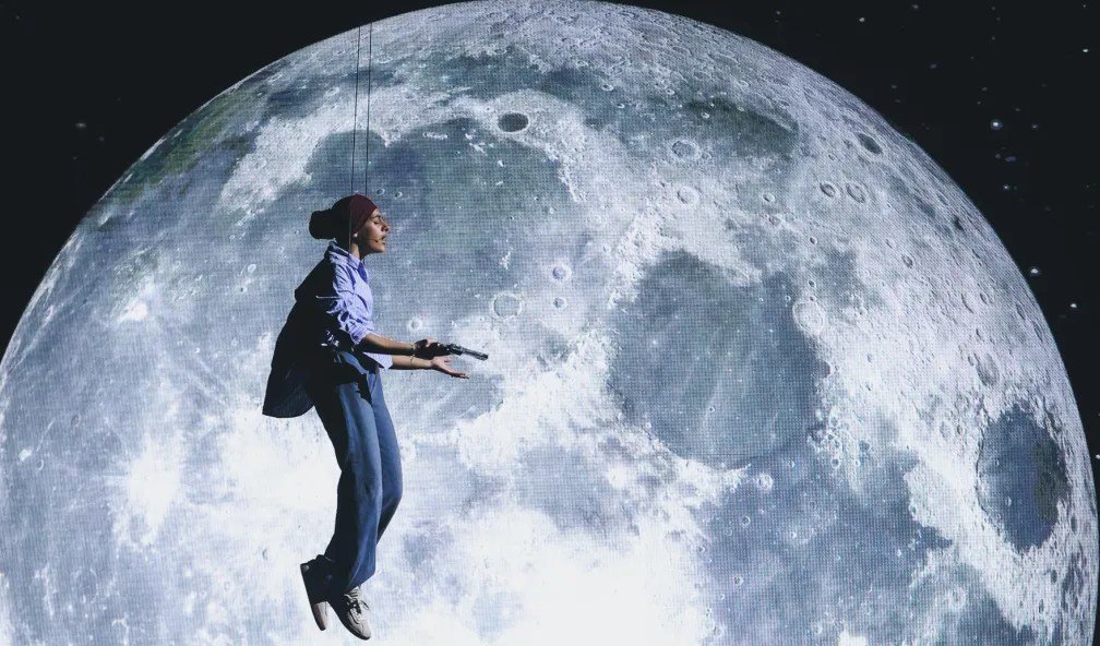 A person in a blue shirt, jeans, and beanie is suspended by wires holding a microphone in front of a large screen displaying the moon.