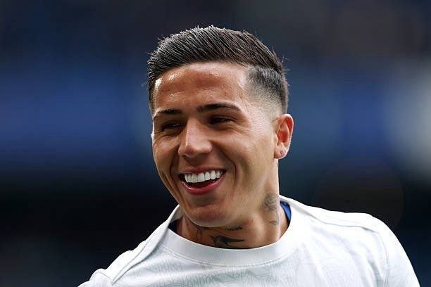 A young man with short, faded hair and neck tattoos smiles broadly while wearing a white shirt against a blurred dark background.
