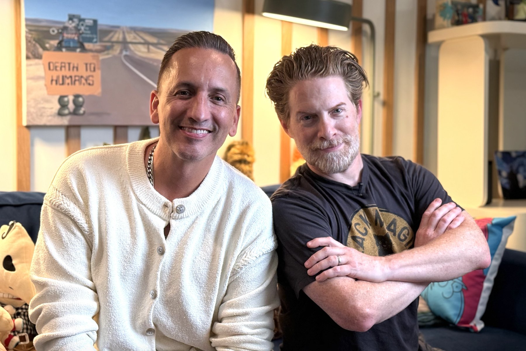 Two men smile while seated on a couch, one in a cream cardigan and the other in a black t-shirt with arms crossed, in front of a poster reading "DEATH TO HUMANS."