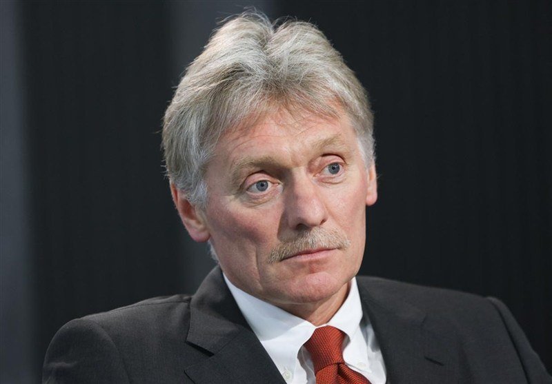 A gray-haired man with a mustache wearing a dark suit, white shirt, and red tie looks off-camera against a dark background.
