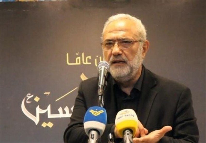 A gray-bearded man in a black suit speaks into multiple microphones, gesturing with his hand in front of a dark backdrop featuring Persian script.