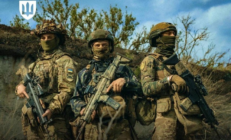 Three camouflaged soldiers wearing helmets and face coverings hold rifles outdoors against a backdrop of bare trees and blue sky.