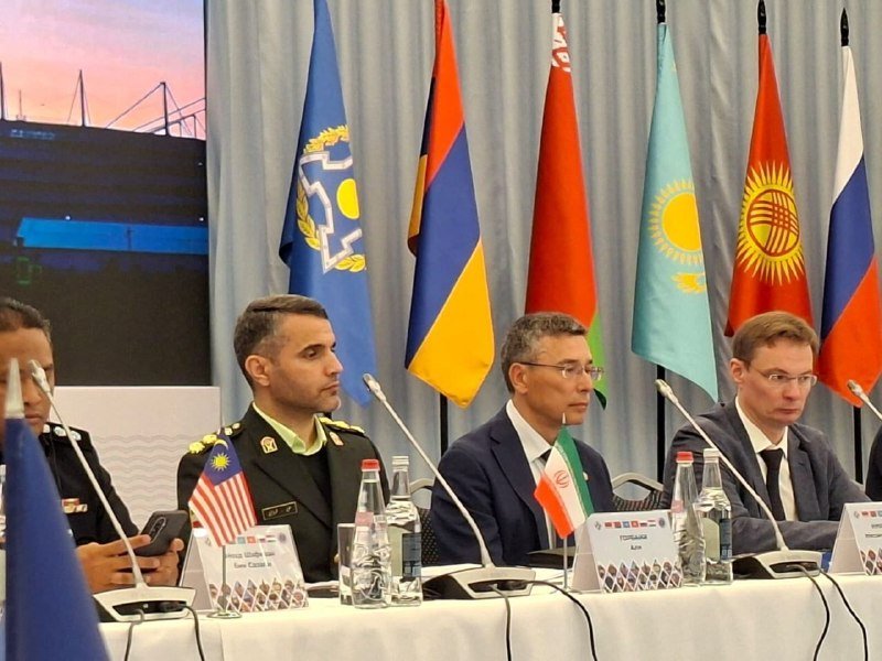 Four men sit at a conference table with microphones and small flags, backed by larger national flags and a stadium backdrop on a screen.