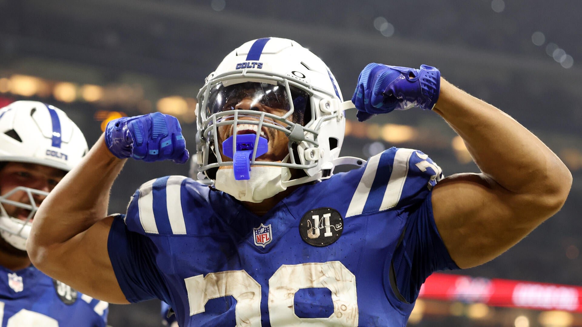 An Indianapolis Colts football player in a blue jersey and white helmet flexes both arms in celebration, with a teammate visible behind him in a stadium setting.