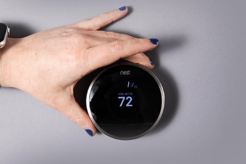 A hand with blue nail polish adjusts a black Nest thermostat displaying "COOL SET TO 72" on a gray wall.