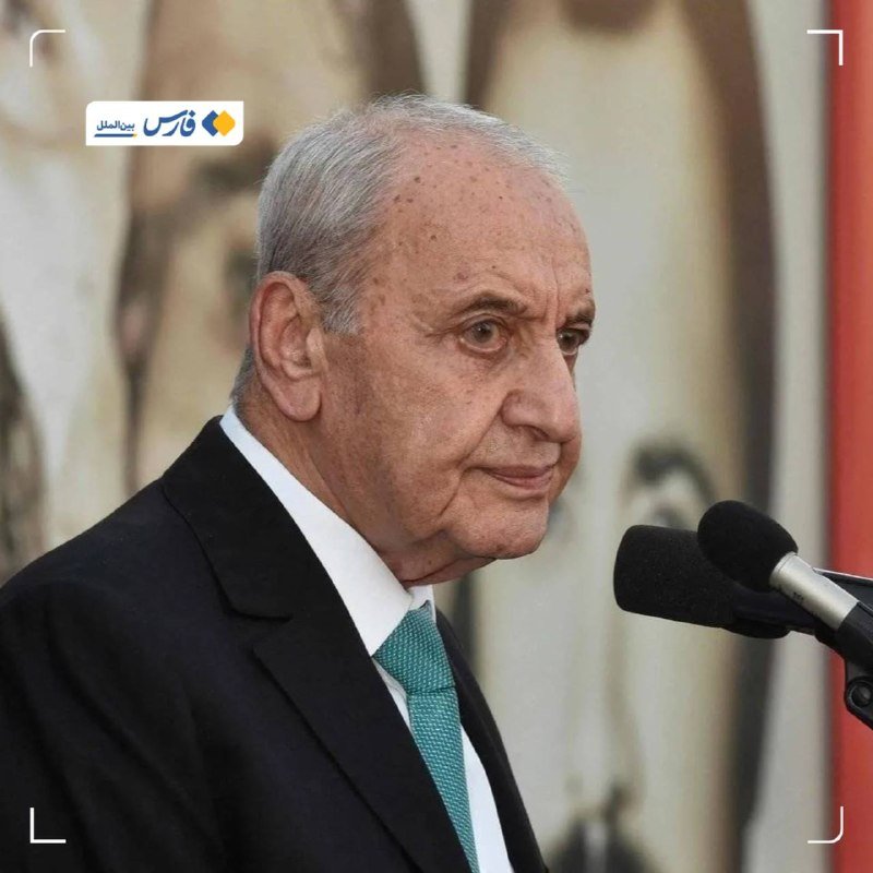 An elderly man with gray hair, wearing a black suit and teal tie, speaks at a microphone against a blurred background.