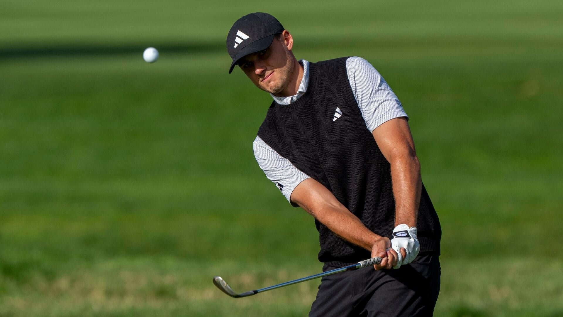 A golfer in a black Adidas cap and sweater vest chips a ball onto a green fairway.