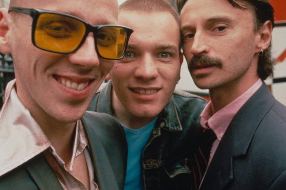 Three men pose closely together for a close-up photo: one on the left wears yellow-tinted sunglasses, the center man sports a shaved head, and the right man has dark hair and a mustache.