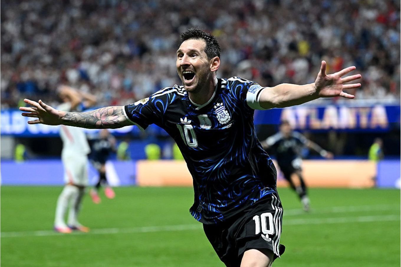 A soccer player wearing a black and blue jersey with the number 10 celebrates with arms outstretched on a field before a blurred crowd.