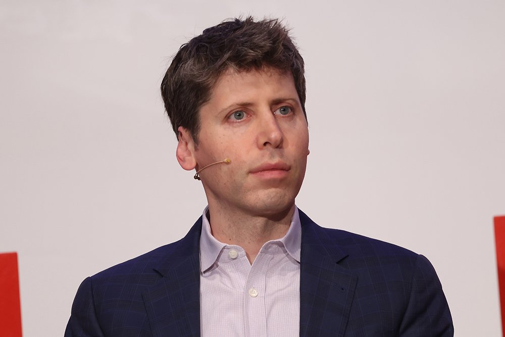 A man with dark hair wearing a dark blazer and light button-down shirt speaks into a headset microphone against a light background.
