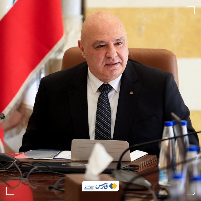 A bald man in a black suit and dotted tie speaks while seated at a desk with a tablet, papers, water bottles, and a red flag behind him.