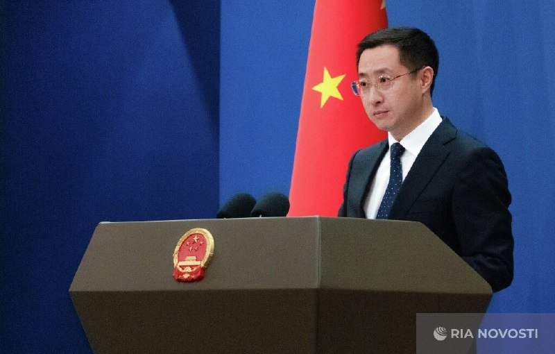 A man in a dark suit and tie speaks at a podium bearing a national emblem, with a red flag featuring yellow stars behind him.