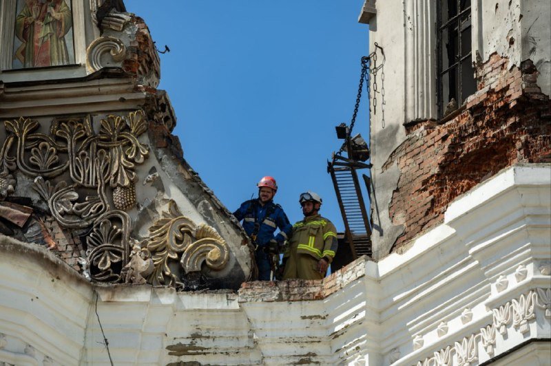 Two rescue workers in helmets and protective gear stand atop the damaged, ornate facade of a partially collapsed building against a clear blue sky.