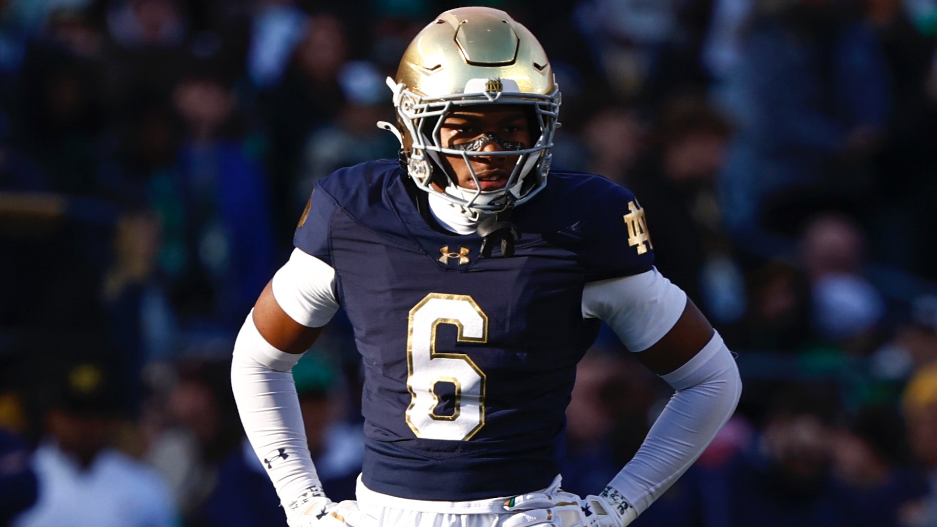 A football player wearing a navy blue jersey with the number 6 and a gold helmet stands on the field during a game.