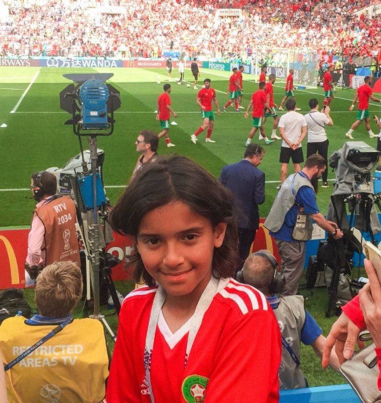 A young person in a red jersey stands in the foreground at a crowded soccer stadium, with players in red and green warming up on the pitch behind broadcast cameras and staff.