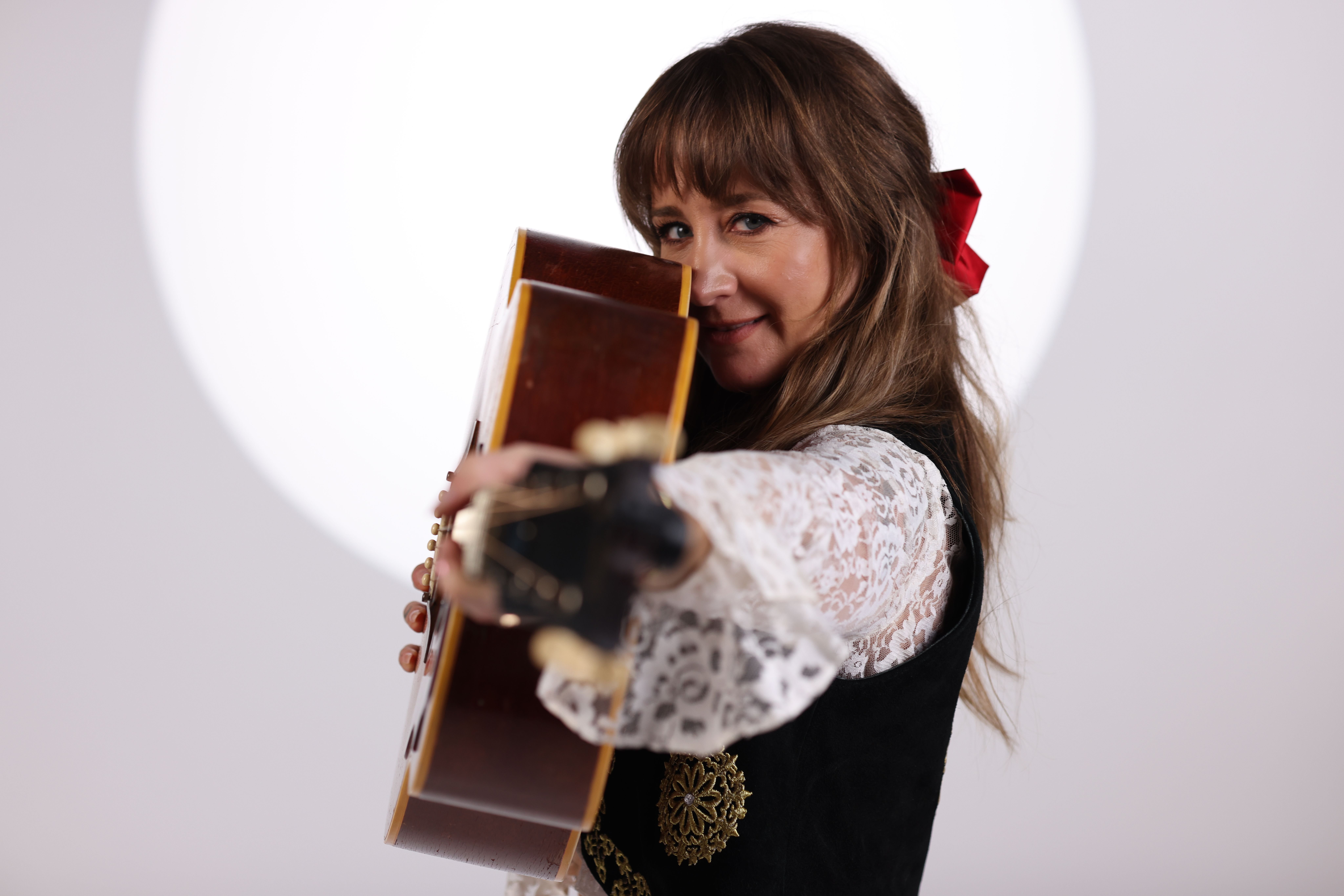 A woman with a red hair bow holds an acoustic guitar against a black vest with gold embroidery and white lace sleeves.