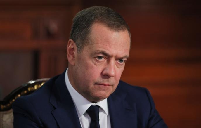 A man in a dark suit and tie sits indoors against a wood-paneled background, looking downward with a serious expression.