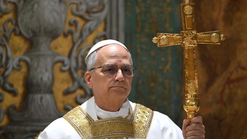 A Catholic clergyman wearing a white skullcap and gold-embroidered vestments holds an ornate golden processional cross.