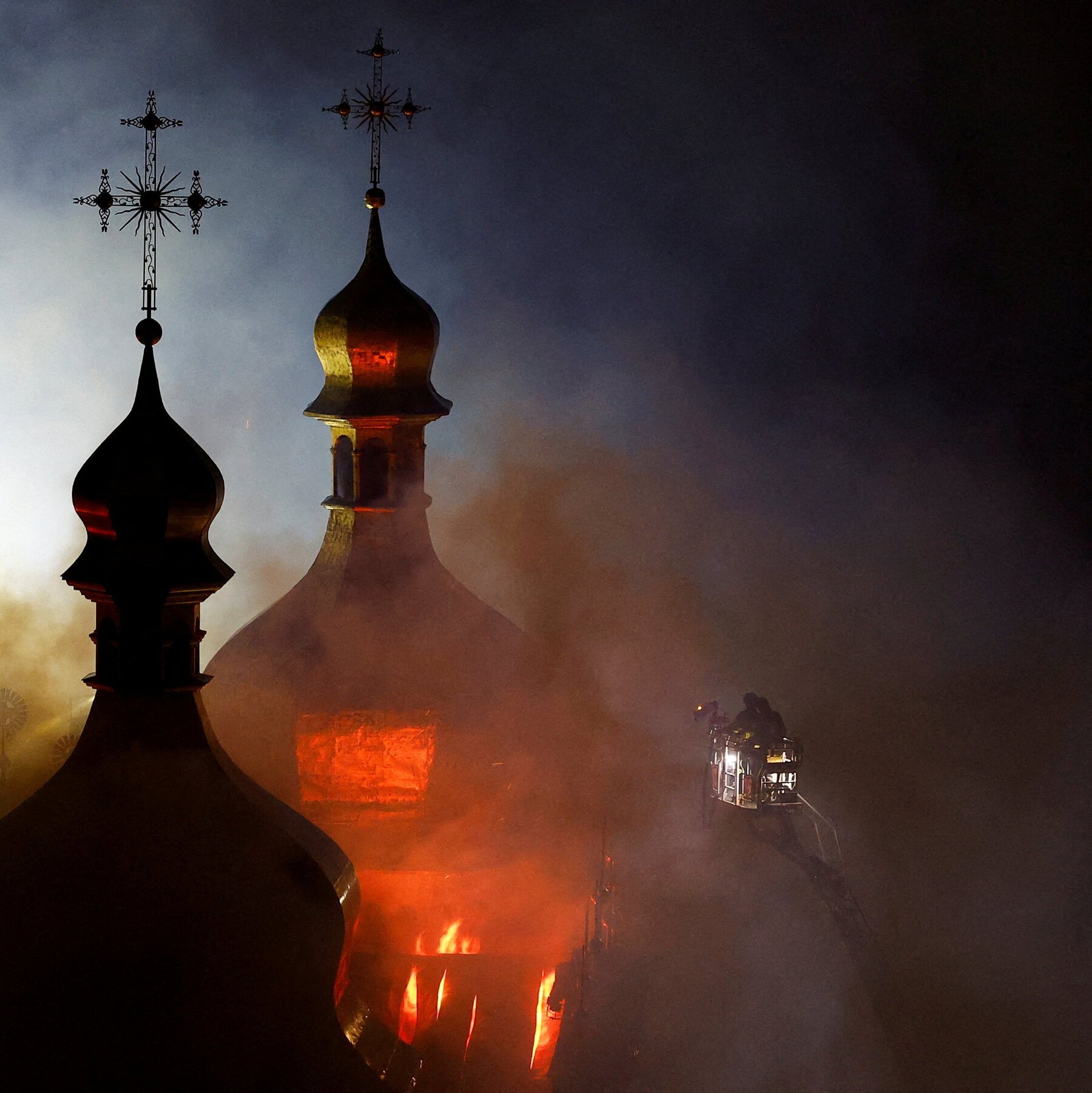 Flames engulf an Orthodox church with onion domes and crosses at night, as firefighters in an elevated platform battle the blaze amid thick smoke.